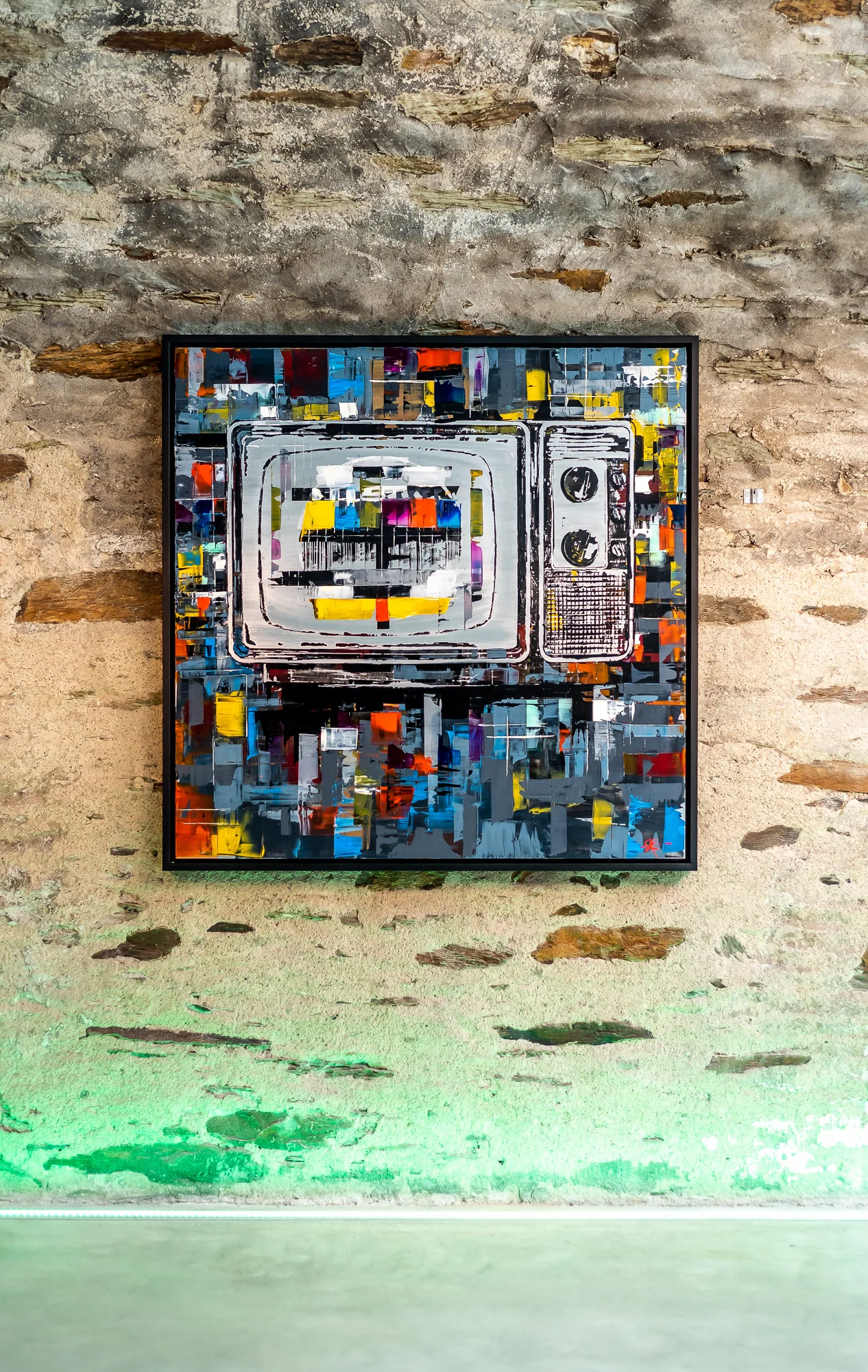 Test Card TV, an original mixed media painting by Paul Kenton, hangs in a black frame on a rough stone wall. A vintage television is drawn in white and grey at the centre, surrounded by bold blocks of yellow, orange, red and blue.
