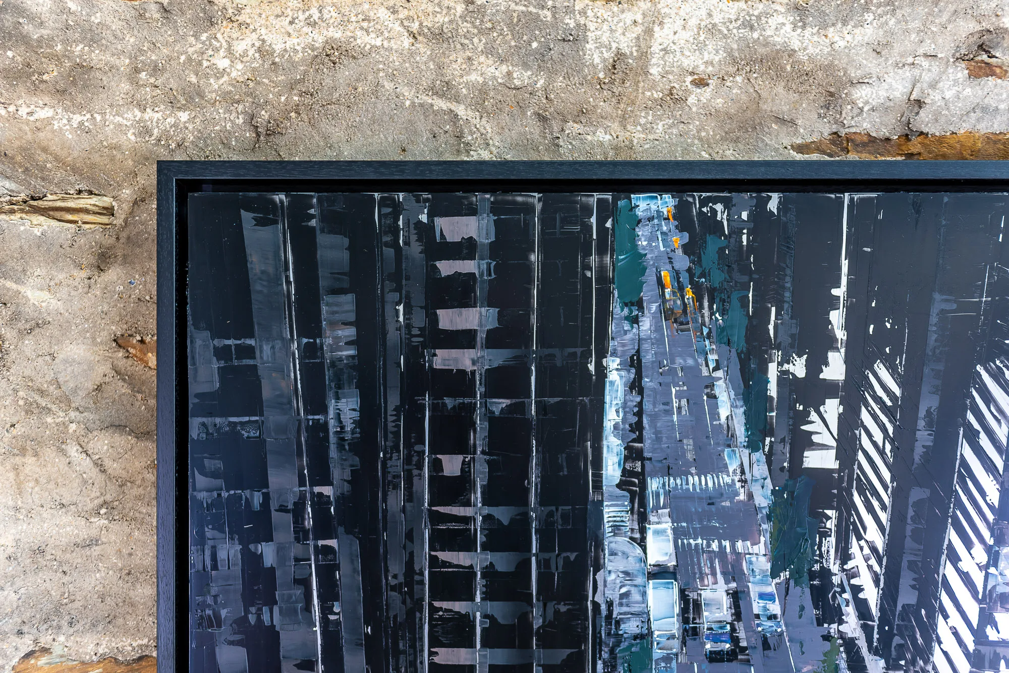Close-up of the top corner of Towering Perspective, an original painting by Paul Kenton, showing a dark city street canyon with bold, scraped white and grey strokes and yellow taxis below, in a black frame against a weathered stone wall.