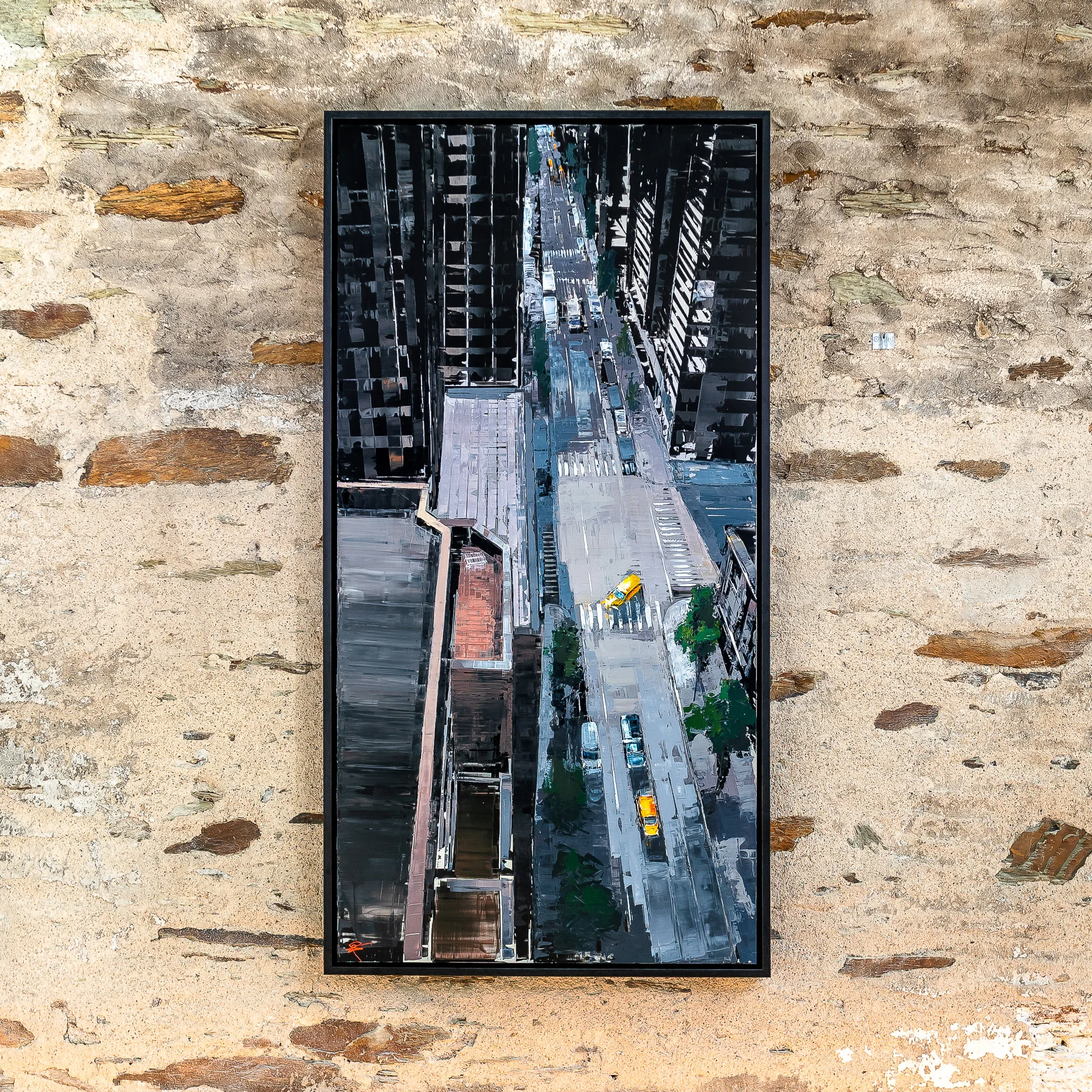 Towering Perspective, an original painting by Paul Kenton, hangs in a black frame on a rough stone wall. It shows a high aerial view down a narrow New York street between dark skyscrapers, with yellow taxis and a green-tinged palette.