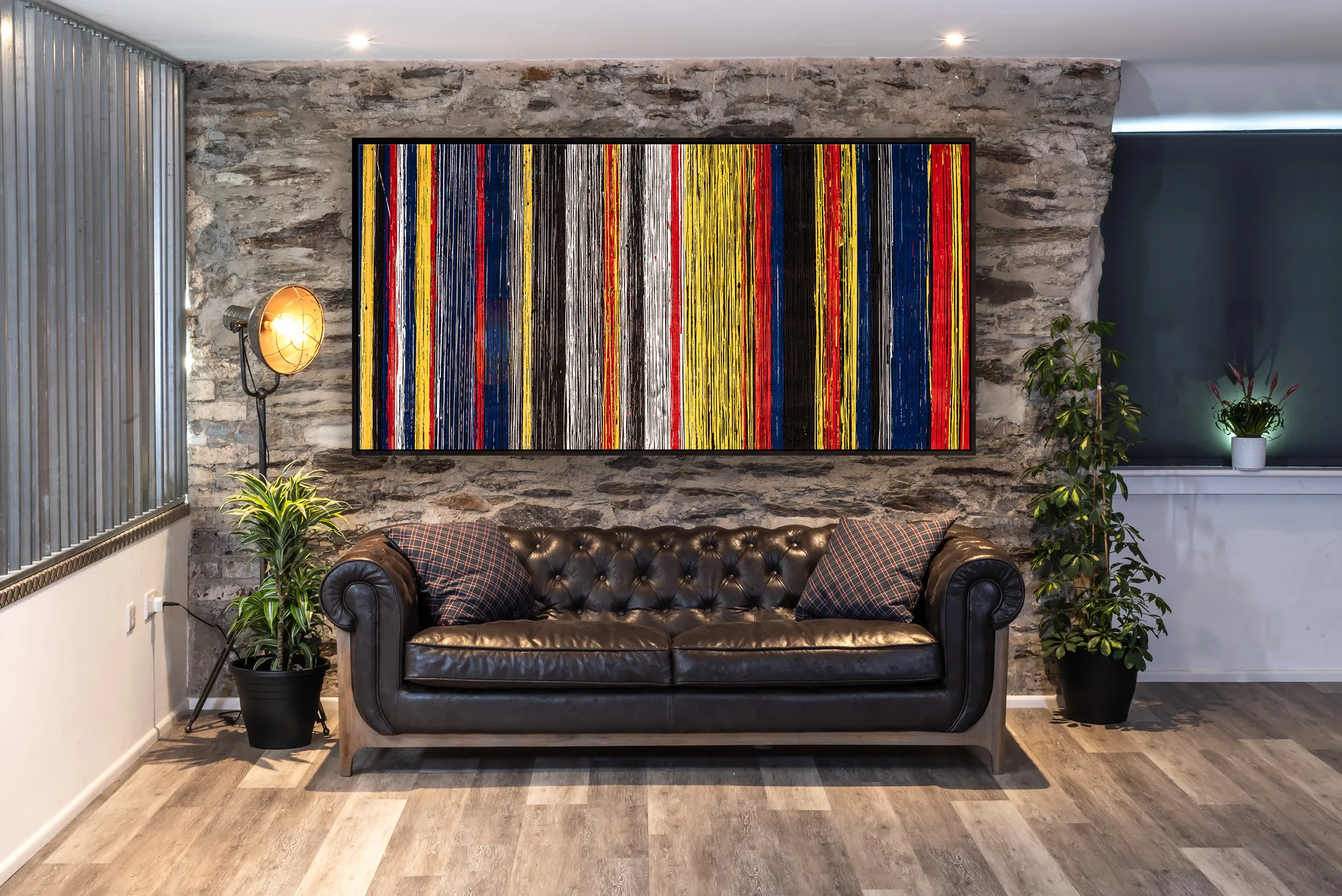 Uniform Oscillation, an original acrylic painting by Paul Kenton, hangs above a brown leather Chesterfield sofa against a stone wall, its vertical bands of yellow, red, blue, black and white bright against the room.