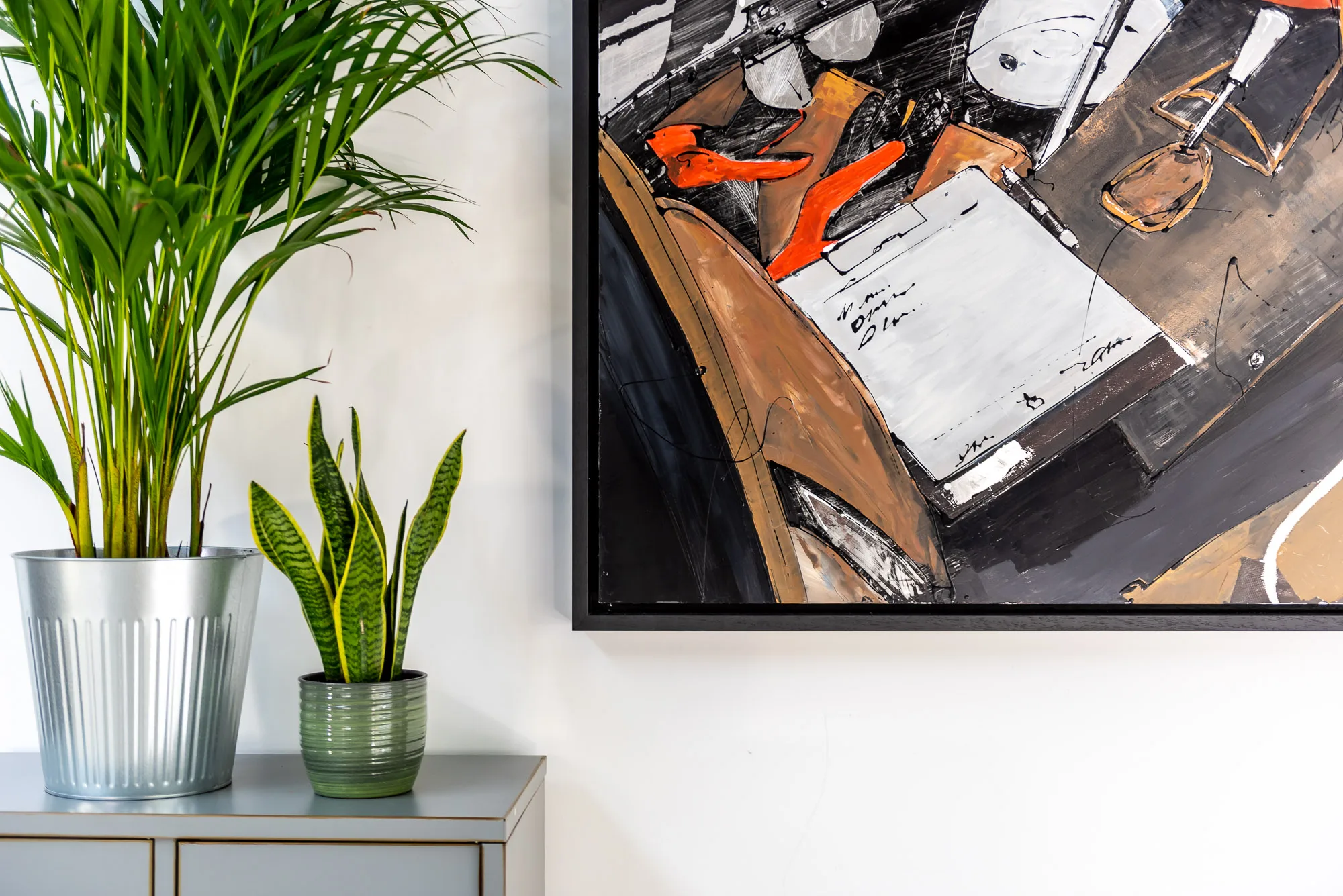 Start with the Checklist, an original mixed media painting on aluminium by Paul Kenton, seen partly cropped on a white wall. Bold orange heels, black and white sketched forms and warm brown tones fill the canvas, beside potted plants on a grey sideboard.